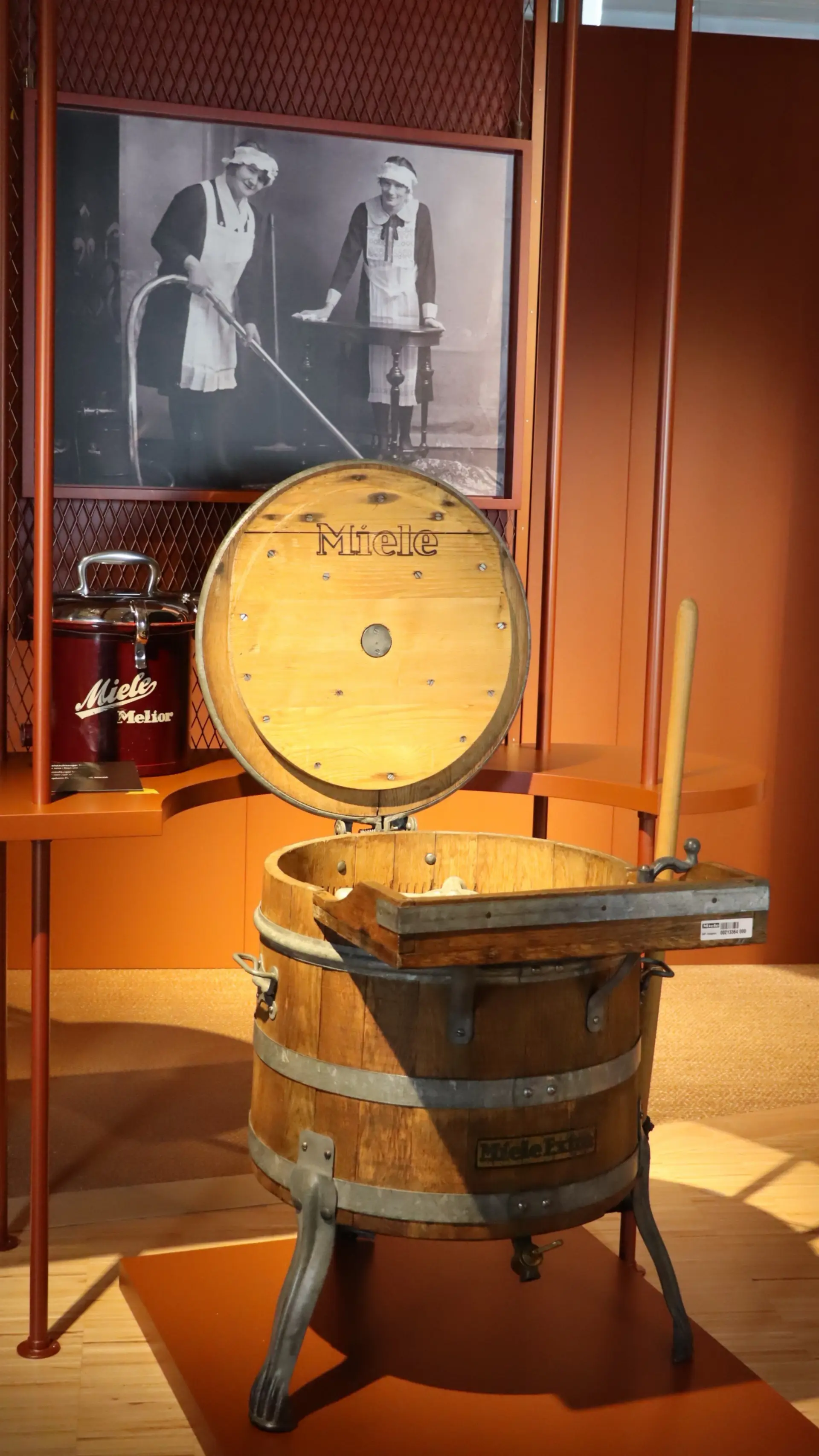 Eine runde Holzwaschmaschine mit geöffnetem Deckel steht vor einer Wand mit einem großem Foto