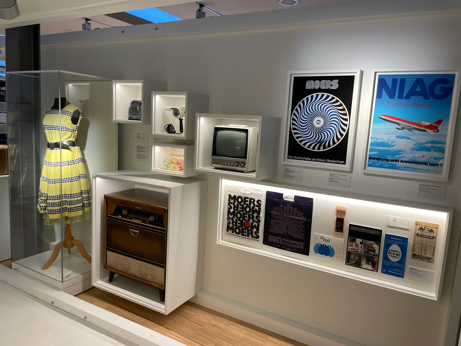 Verschieden große Fächer, in denen unterschiedliche Gegenstände untergebracht sind. Seitlich daneben steht eine Kleiderpuppte mit einem gelben Kleid